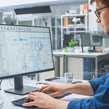 Over the Shoulder Shot of Engineer Working with CAD Software on Desktop Computer, Screen Shows Technical Drafts and Drawings. In the Background Engineering Facility Specialising on Industrial Design Vescient Engineering